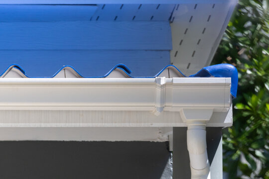 Close-up of white rain gutter under blue tiled roof with greenery