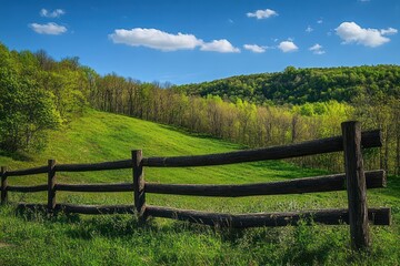 Fototapeta premium sunlit wooden fence bordering rolling green hills and leafy forest under a blue sky with fluffy clouds, evoking peaceful rural tranquility