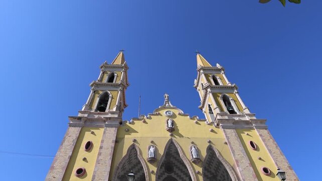 View of the yellow facade and twin bell towers of the Catedral Basilica de la Inmaculada Concepcion under a clear blue sky in Mazatlan, Sinaloa, Mexico.