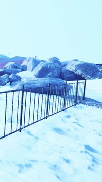 A metal fence stands in front of large rocks surrounded by snow. The setting shows a cold environment with a clear sky. This scene reflects winter conditions in a rural area.