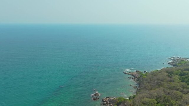 Aerial view of a dense, vibrant green coastal jungle meeting dark blue ocean water. A thin, rocky beach and white foamy waves separate the rugged terrain from the sea.concept	