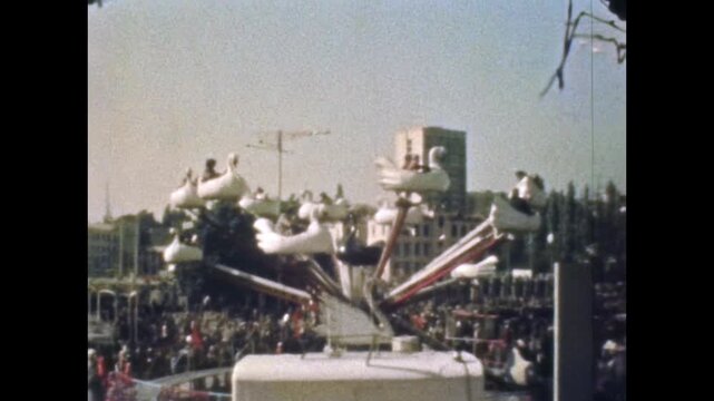 Families and children enjoy a spinning swan ride at a busy amusement park. Colorful outdoor attraction captures joy, holiday leisure fun at fairground. Old film. Archival retro 1960s. Vintage archive