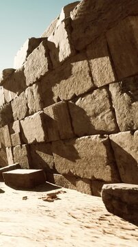 Visitors walk near an old stone wall at an archaeological site under clear blue sky. The sun creates shadows on the stones, highlighting their age and texture.