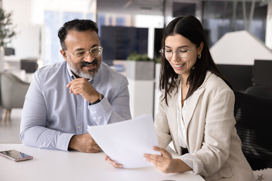 Two multiethnic young and mature teammates reviewing business report, analyzing project results, discussing strategy or commercial proposal seated at desk in modern office. Client services, teamwork