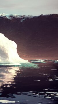 An iceberg drifts in the water near mountains as the sun sets. The sky shows colors of dusk while the water reflects the iceberg and the landscape, creating a scenic view.