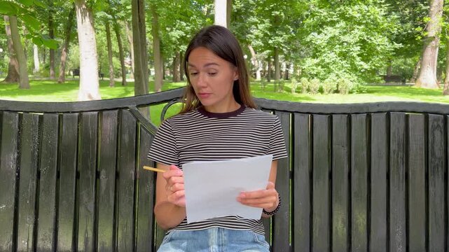 Young woman sitting on a park bench reading a document and writing notes with a pencil. Reviewing important notes in a peaceful outdoor setting.