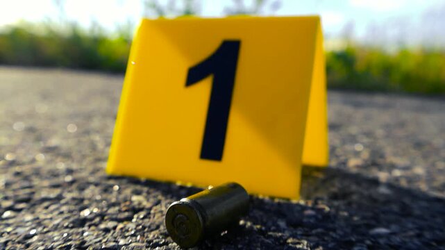 Yellow evidence marker with the number one next to a bullet casing lying on an asphalt road at a crime scene, showing the aftermath of a violent incident and forensic investigation