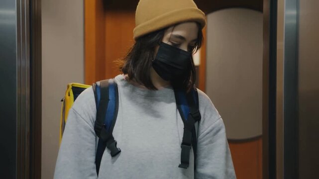 Masked female courier riding elevator with backpack during apartment food delivery shift