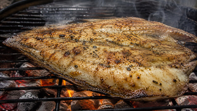 Grilled fish fillet cooking on barbecue grill over hot charcoal with smoke background.