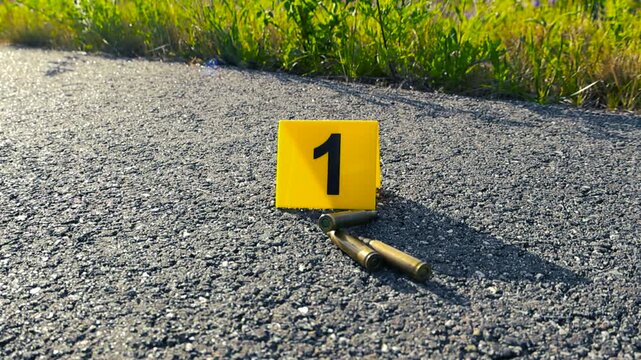 Yellow forensic marker indicating several rifle bullet casings lying on the asphalt road as crucial evidence from a recent shooting, highlighting the seriousness of the crime scene