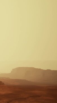 In a desert landscape, large rock formations are visible in the distance. The sky appears hazy with a warm color. The ground is dry and rocky, typical of desert terrain.