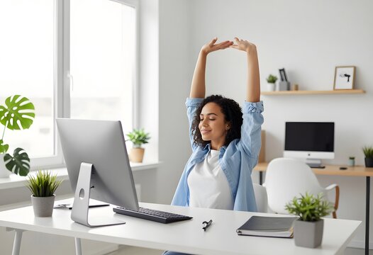 Young woman stretches her arms above her head at a clean desk with a computer monitor, taking a refreshing break from work in a bright home office environment.