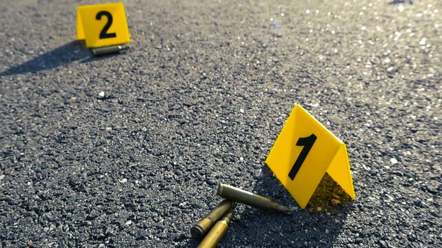 Yellow evidence markers indicating the location of rifle bullet casings on an asphalt road at a crime scene, symbolizing a police investigation into a shooting or violent act with firearms