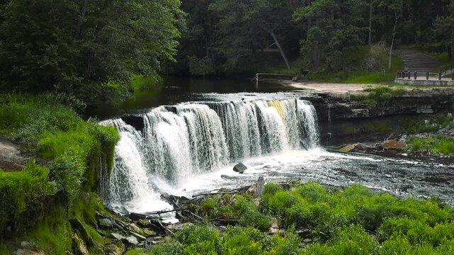 Beautiful view of the Keila waterfall, or Joa, flowing down a limestone cliff surrounded by a lush green forest during a summer day in Estonia, with a small viewing platform on the side