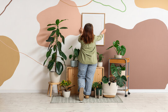 Woman hanging picture frame on colorful wall with houseplants