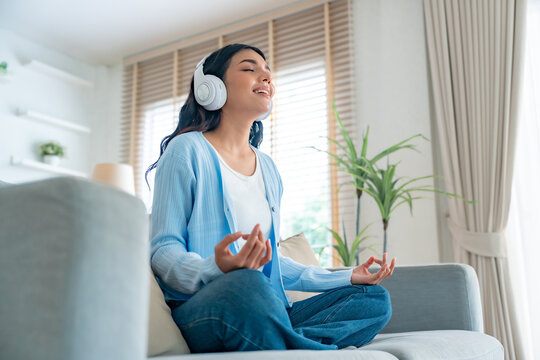 Happy young Asian woman practicing yoga and meditation at home sitting on couch in living room in lotus position and relaxing with closed eyes. Mindful meditation and wellbeing concept