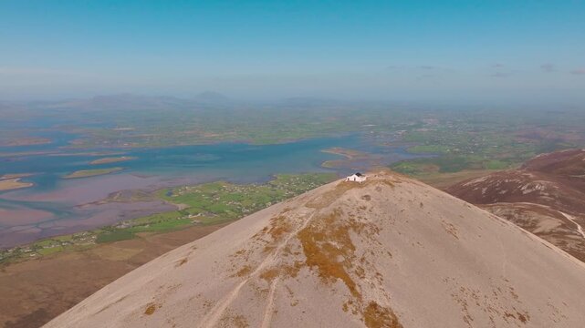 Dramatic drone shot of the Croagh Patrick mountain summit in County Mayo, Ireland. Cinematic view of the holy peak, the small chapel, and pilgrims on the steep trail with Clew Bay in the background.