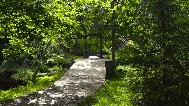 Picturesque stone pathway winding through a sunlit green forest, leading to an old wooden suspension bridge over a small river in a peaceful, natural park setting on a beautiful summer day