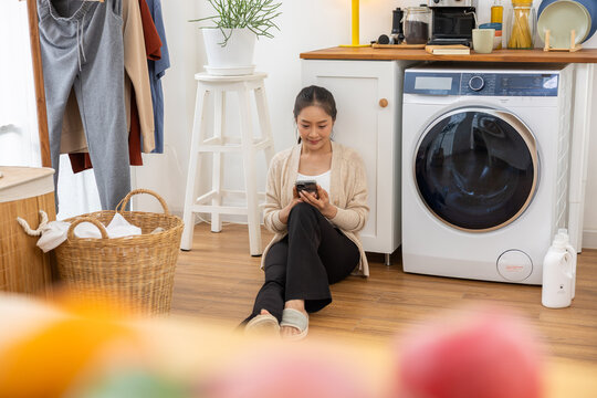 Woman laundry smartphone washer basket clothing home utility room calm woman using smartphone floor bright laundry room near washing machine, wicker basket, hanging clothing, plant, towel
