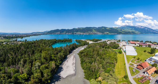 Aerial view of Lake Thun with the Kander river flowing into the lake, surrounded by forests, fields, and mountains under a clear blue sky, Spiez, Canton of Bern, Switzerland.