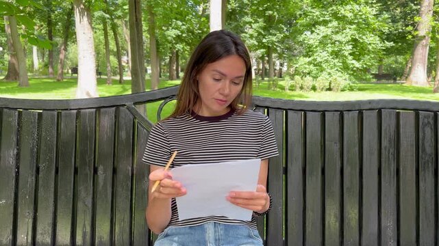 A young brunette woman is preparing for a test sitting on a bench in a city park. working in a peaceful natural environment.