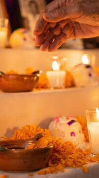 Hands placing marigold petals on a traditional Mexican Day of the Dead altar with candles and sugar skulls, Dia de los Muertos ofrenda at night.