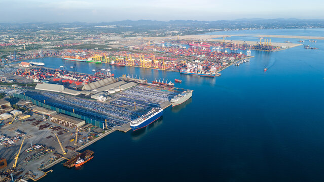 roll-on roll-off (RoRo) vessel docks at a major industrial port facility, surrounded by thousands of new vehicles staged for global export alongside busy container shipping terminals.