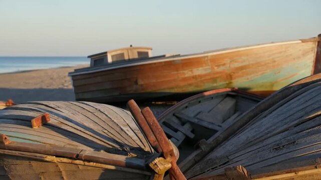 Panning shot of weathered wooden boats resting on pebbled beach shoreline with calm sea and low morning light
