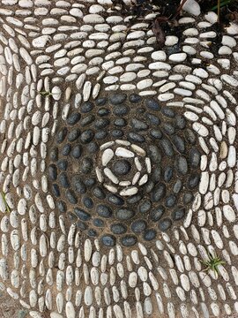 stone wall background. Concentric pebble mosaic pattern with alternating dark and light stones, creating a decorative spiral design for outdoor flooring