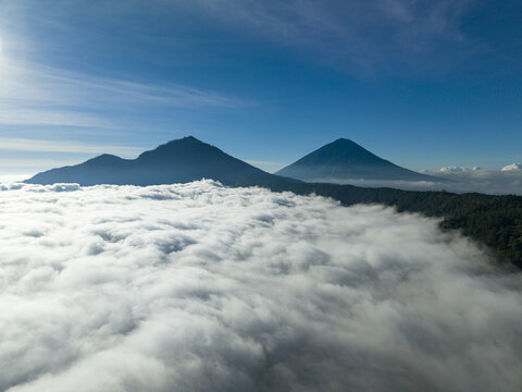 Aerial view of Mount Batur and Mount Abang rising above a thick sea of clouds under a clear blue sky in Kintamani, Bali, Indonesia.