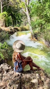 Young female tourist with straw hat and backpack sitting on rocks looking at rushing river in green forest