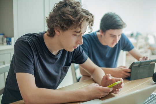 Two teenage boys using smartphone and tablet on table at home