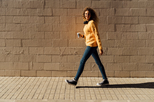 Young woman holding disposable cup while walking on footpath by brick wall during sunny day