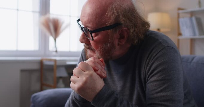 Sad elderly man in glasses sitting on couch, clasping hands and looking down in sorrow or deep contemplation. Close up portrait of unhappy senior male feeling deep loneliness, grief and regrets.