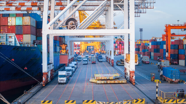 Container trucks driving under massive gantry cranes at a busy commercial port terminal, supporting global maritime logistics, sea freight supply chain, and international trade operations.