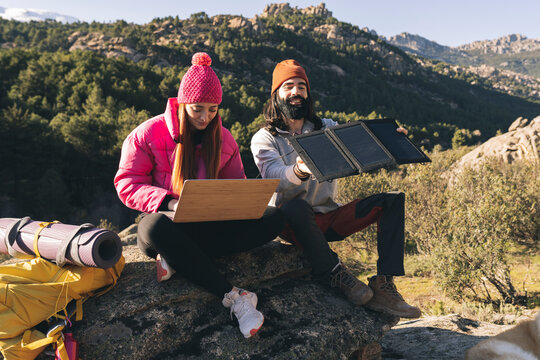 Young woman using laptop sitting by man adjusting solar panel on sunny day