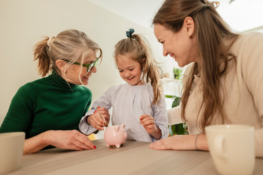 Smiling mother and grandmother teach daughter saving money indoors