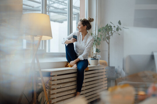 Smiling woman holding mobile phone sitting on widow sill at home