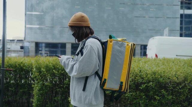 Side view of delivery woman using phone near hedge and modern skyline