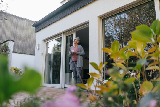 Mature woman with cup of coffee standing at terrace door