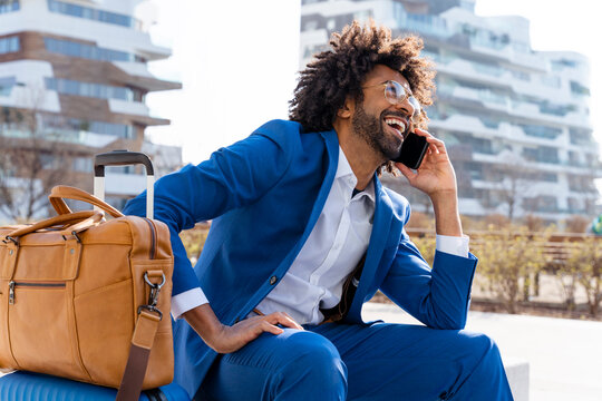 Happy businessman with suitcase and shoulder bag talking on smart phone sitting on concrete block