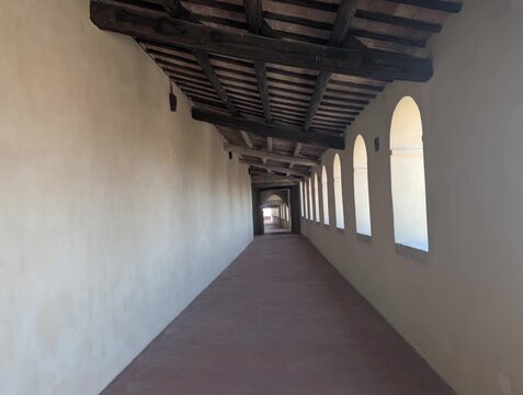 Corridoio porticato con travi in legno e pavimento in cotto, illuminato da archi laterali che creano giochi di luce e ombra. Atmosfera storica