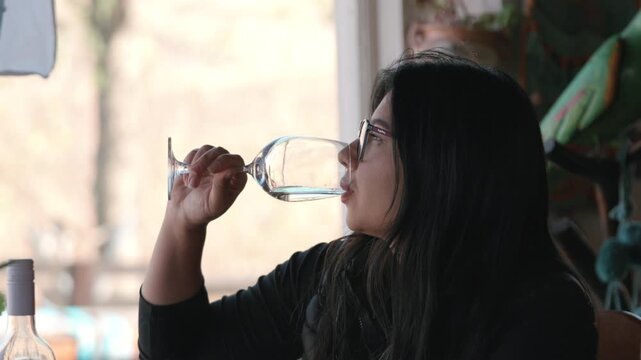 Woman with eyeglasses at a rustic mountain lodge restaurant, picking up a glass of white wine to take a sip. Static closeup profile shot.