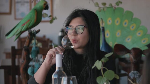 Woman with eyeglasses sitting at a rustic restaurant table, swirling and tasting a glass of regional white wine with wooden sculptures background. Static closeup.