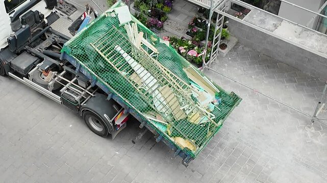 Construction debris removal truck with scaffolding and plants on pavement. A truck is parked on a paved surface, its dumpster filled with construction debris and covered by a green net, with