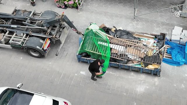 Worker secures debris in a dumpster with a green net for transport. A man in work clothes carefully adjusts a green safety net over a large dumpster filled with various waste materials and debris.