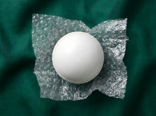 White cricket ball resting on bubble wrap sheet placed atop dark green draped textile texture for sports supply logistics