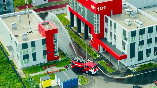 Red fire truck sprays a high water stream from a hose against a firehouse building facade. Firefighter crew background training exercise drill layout in a modern safety department station facility.
