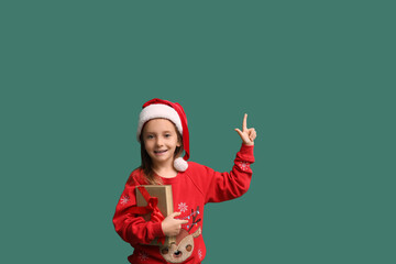 Obraz premium Cute little girl in Santa hat with gift book pointing at something on green background