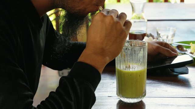 Bearded man drinking fresh green avocado juice using straw while holding smartphone at cafe table.